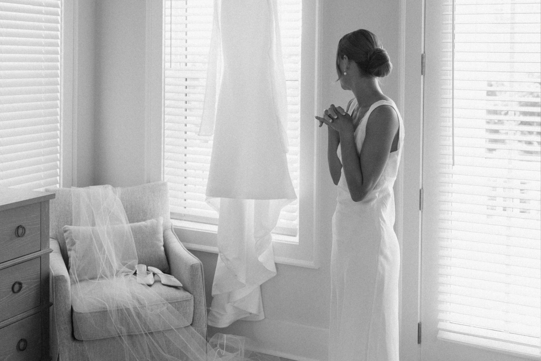Bride standing in her bridal suite wearing a white slip dress with lace details for the morning of her wedding day