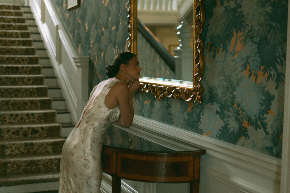 Bride wearing a floral slip dress with lace details putting on earrings in front of a mirror