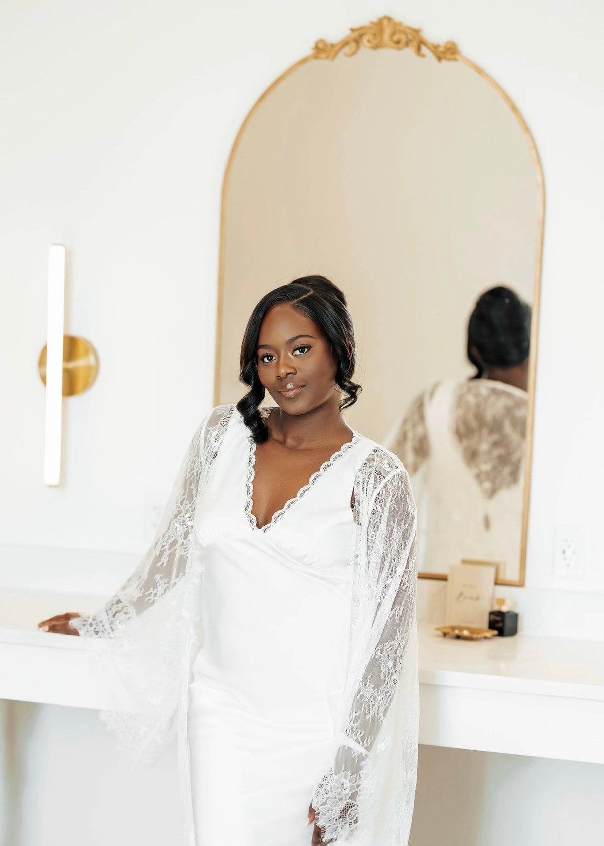 Bride in a white lace slip dress standing in front of a large mirror the morning of her wedding day