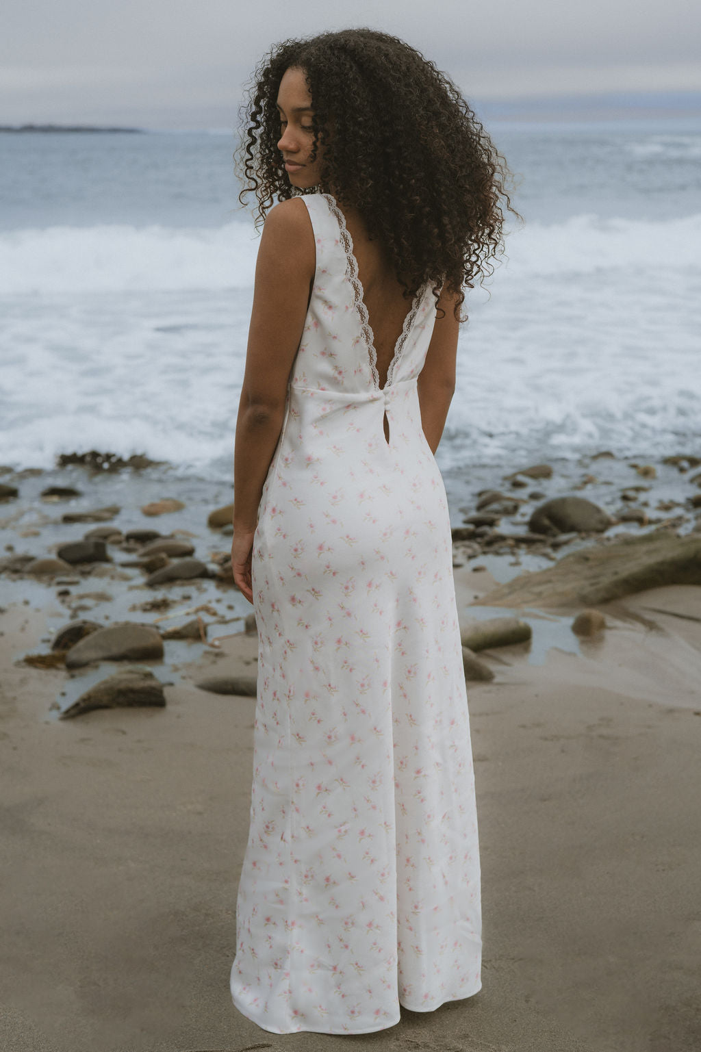 Woman in a white floral slip dress standing on a beach with ocean waves in the background