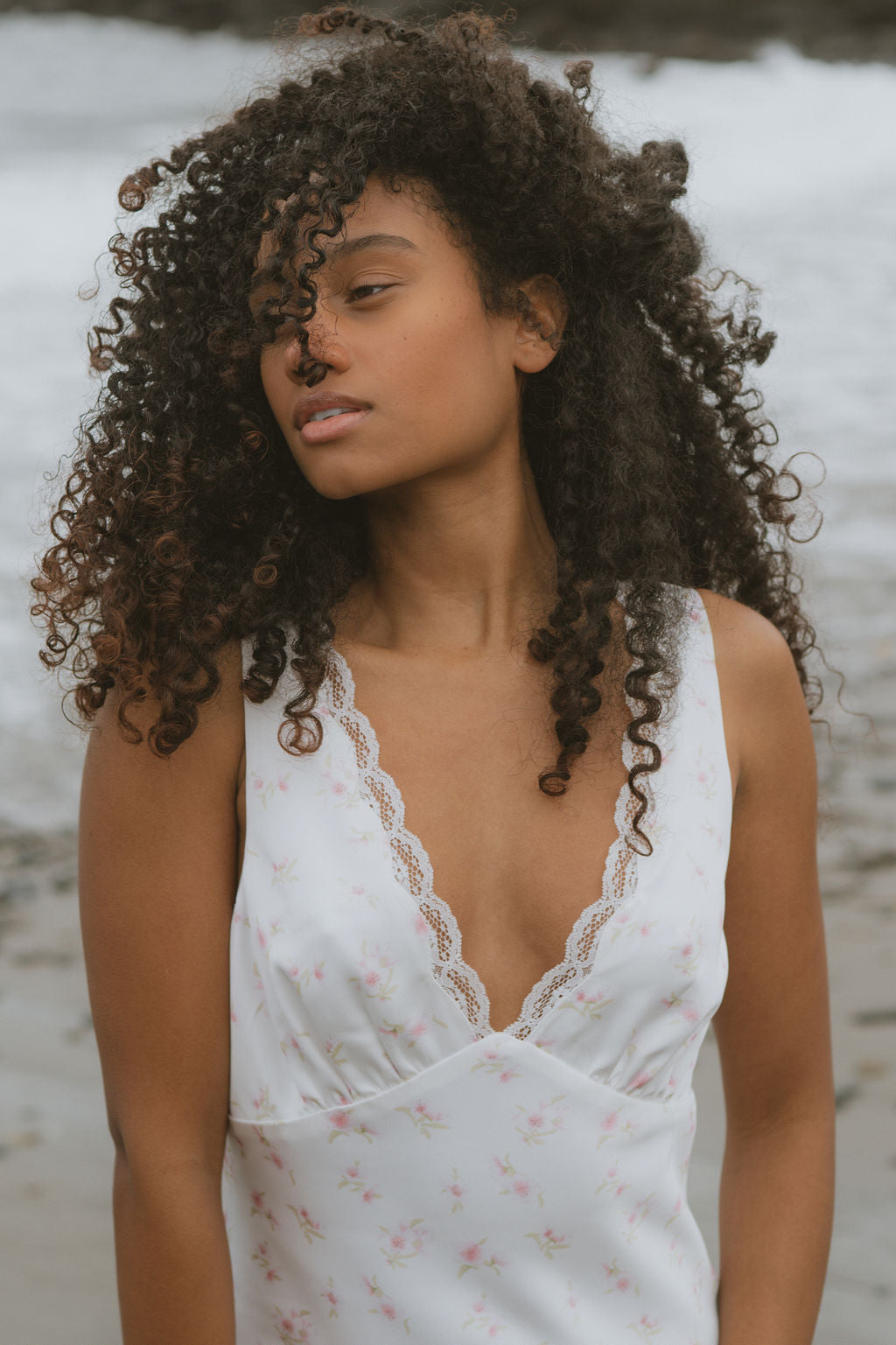 Woman with curly hair wearing a white floral slip dress on a beach.