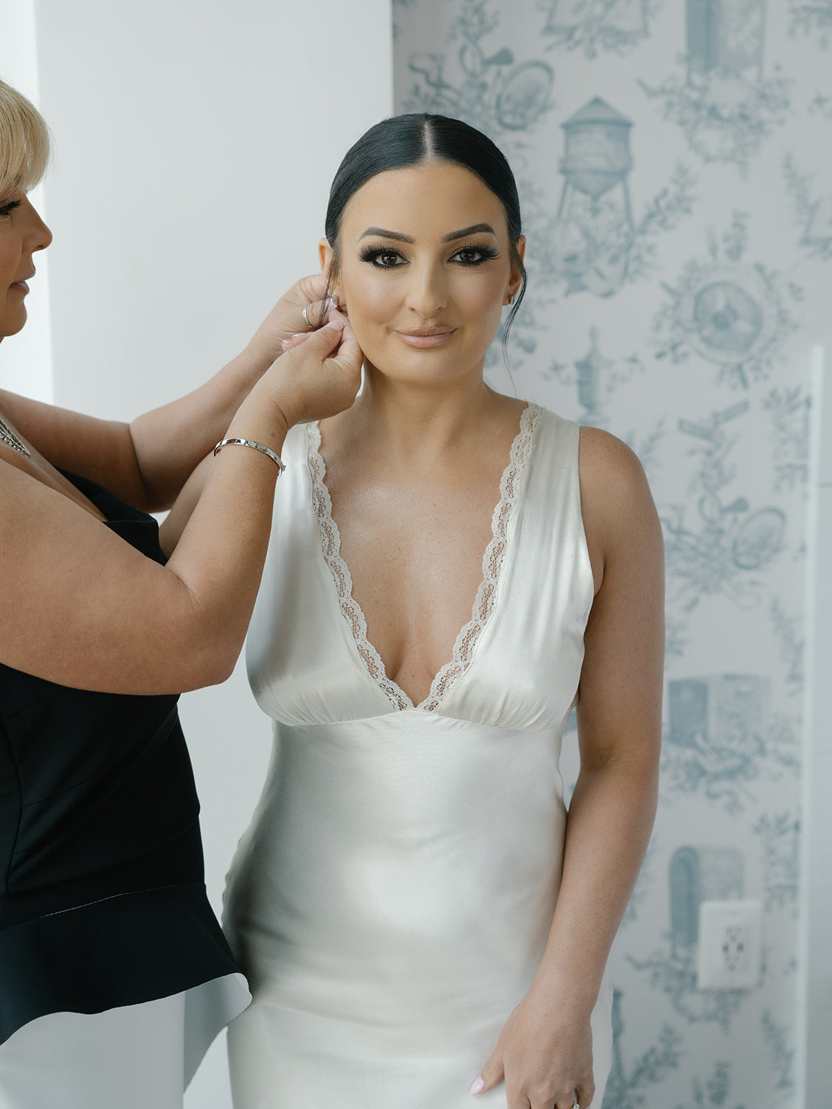 Bride in a white slip dress having an earring adjusted by her mother against a decorative wall.