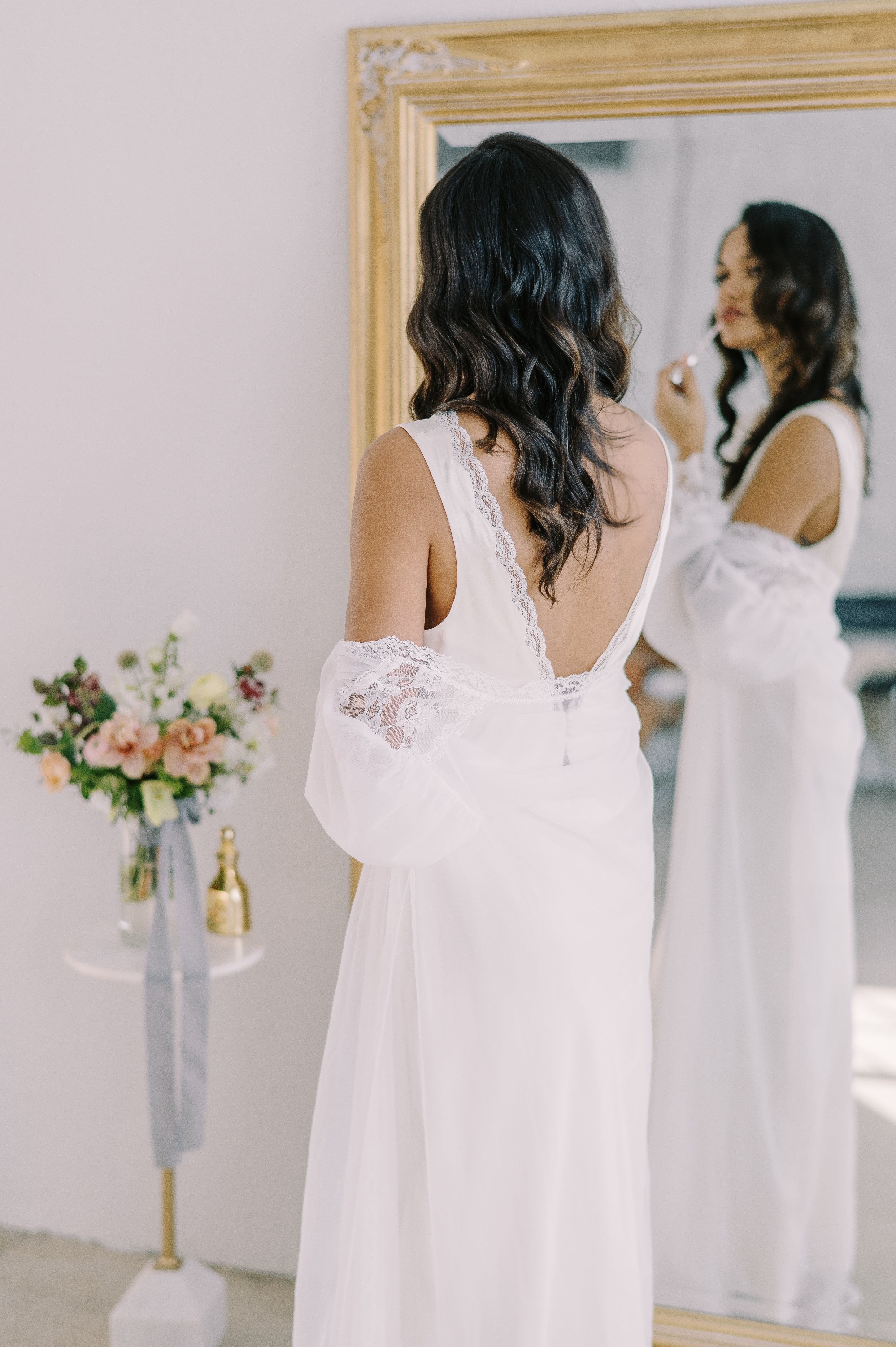 A bride in a white lace-trimmed slip dress applies lipstick in front of a gold-framed mirror. 