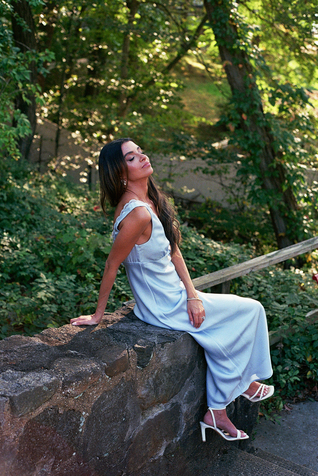 Woman in a something blue bridal dress sitting on a stone wall in a park.