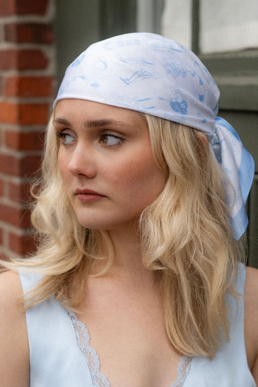 Woman wearing a light blue headscarf with a bridal pattern, standing in front of a brick wall.