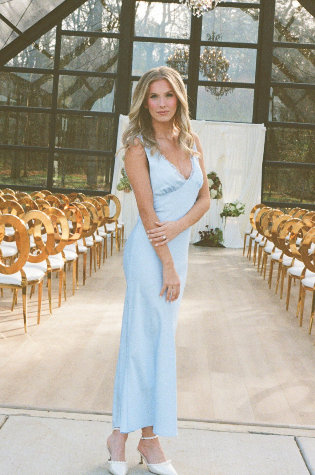 Woman in a light blue dress standing in a large, elegant room with glass walls and wooden chairs.