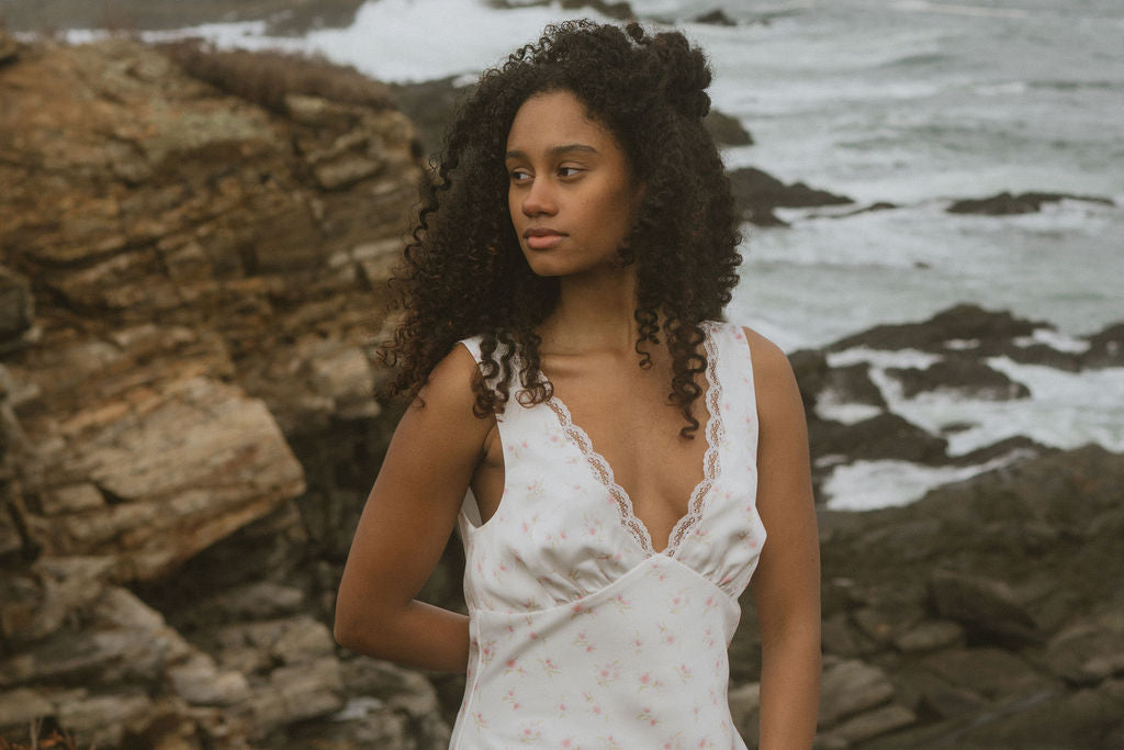 Woman in a white floral slip dress standing on a rocky coastline